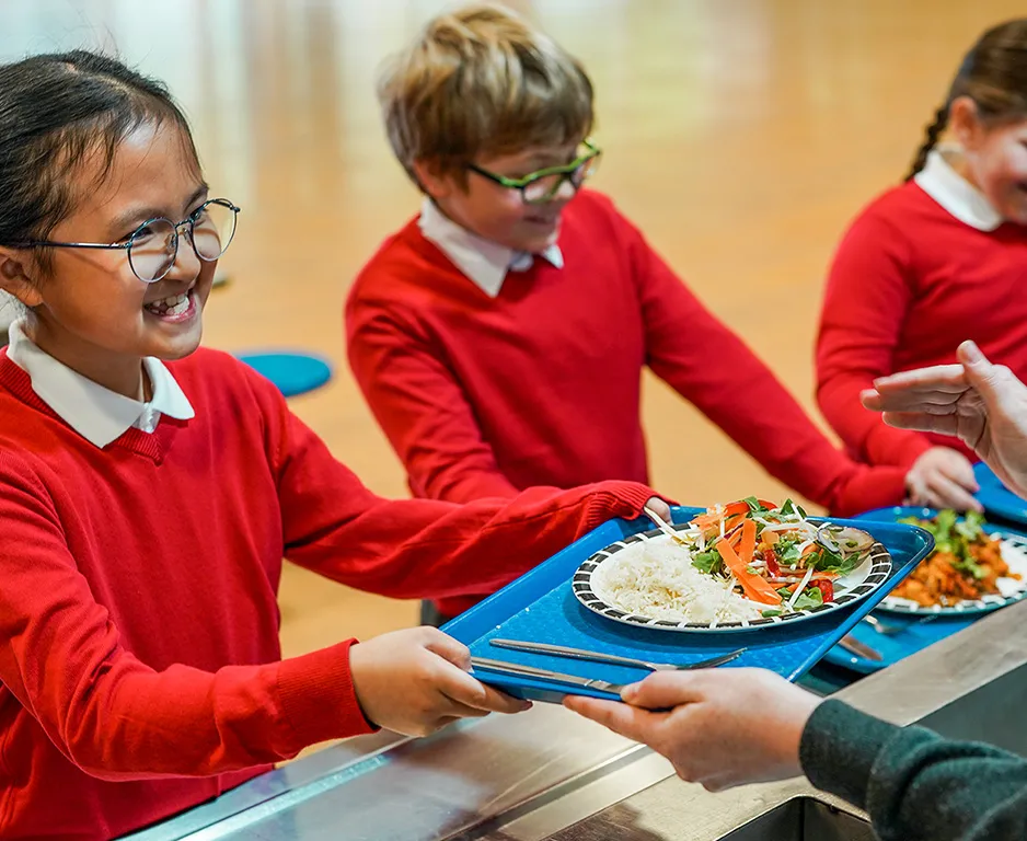 Smiling primary school children being handed lunch trays with a plate of colourful vegetables and rice.